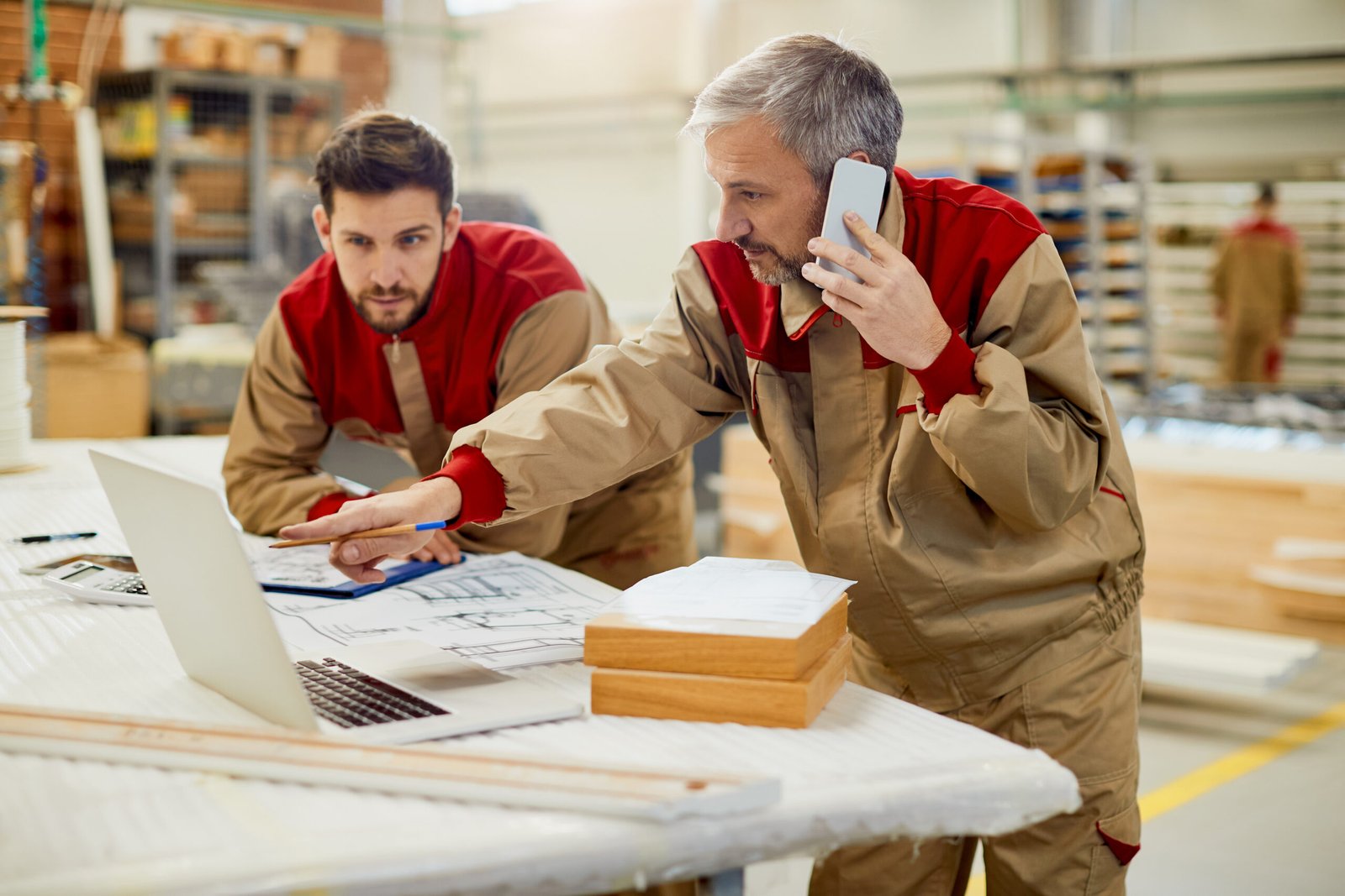 Mid adult worker communicating over mobile phone while using laptop with his colleagues at carpentry workshop.